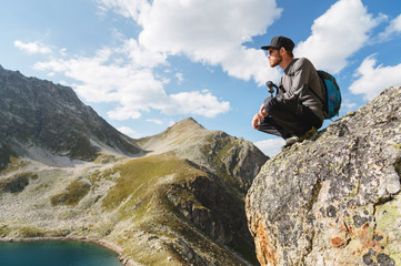 A bearded hipster tourist in sunglasses with a backpack sits on the edge of a cliff high in the mountains near the mountain lake