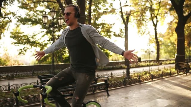 Happy excited young man riding bike in the park and listens to the music in black headphones. Man with outstretched hands. Enjoyment, hanging outdoors in the park. Front view