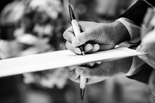 Foto b/n di una mano che firma di un contratto con riflesso su tavolo 