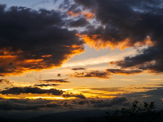 Beautiful sunset with orange and gold tones with large black clouds. Golden dramatic clouds over the horizon of mountains in a rural scene.