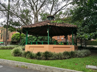 Fototapeta na wymiar Small traditional kiosk or gazebo with green wooden columns and small circular roof with clay tiles. Green area with vegetation and trees.