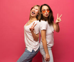 Two female friends hugging and having fun together, showing peace gesture while looking at camera, isolated over pink background.