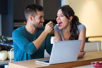 Young handsome man giving yogurt to his beautiful girlfriend in the kitchen at home.