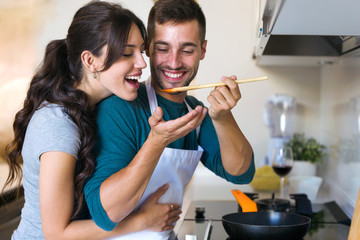 Handsome young man giving his wife to try the food he is preparing in the kitchen at home.