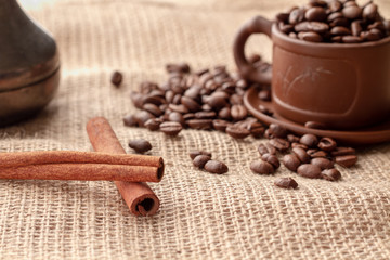 Two sticks of natural cinnamon next to the coffee beans, which are scattered near a brown clay cup...