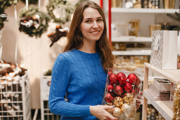 Happy woman with red and gold balls for Christmas tree decor in New Year shop. Brunette smiling girl at christmas market. Shopping. Lifestyle.
