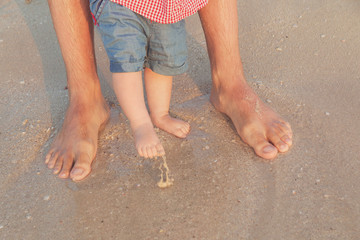 Man and baby feet standing in shallow water waiting for the wave. Bare feet father and his little daughter or son staying in the sand near the sea. Concept of travel and holidays. Toned. Soft focus.