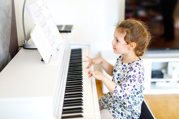 Beautiful little kid girl playing piano in living room or music school. Preschool child having fun with learning to play music instrument. Education, skills concept