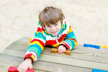 Happy blond kid boy having fun and climbing on outdoor playground. Funny joyful child smiling and making sports. Summer, spring and autumn leisure for active kids. Boy in colorful fashion clothes.