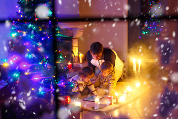 Father and two little toddler boys sitting by chimney, candles and fireplace and looking on fire. Family celebrating Christmas. With Xmas tree and lights on background. Kids happy about gifts