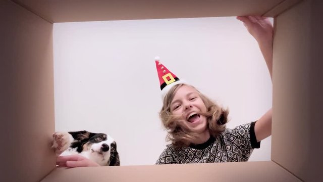 Young happy teen girl with Christmas hat opening cardboard box and looking inside. Child with dog unboxing gift - different versions of emotions. Concept of holidays and New Year.