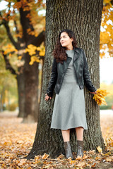 Woman with yellow leaves stand near big tree in autumn city park.