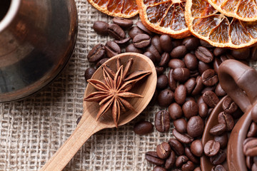 Wooden spoon with anise stars lies between the Turk for making Coffee and coffee, oranges, and overflowing glay Mug which are scattered on the table with burlap cloth. Top view.
