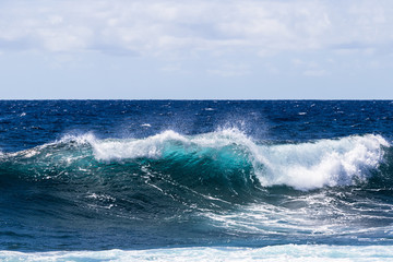 Wave breaking near shore on South Point, on Hawaii's Big Island. Foam on top of wave's clear blue-green water; deep blue Pacific ocean in the background.  