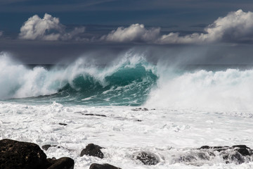 Large wave from tropical storm breaking on western Kona coast in Hawaii. White spray and foam on the water. Rocks in foreground. 
