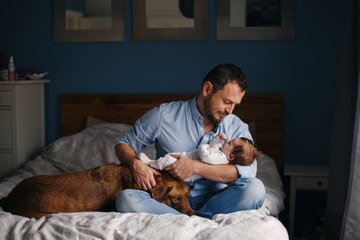 Portrait of middle age Caucasian father with newborn baby. Dog pet laying on bed. Man parent holding child in hands. Authentic lifestyle documenatry moment. Single dad family life. - 233070455