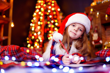 Little charming girl in a plaid dress, fur vest, Santa hat is lying on the floor and writes a letter, draws with a pencil. lights of garlands. warm comfortable room