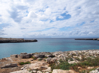 clifftop view of the cove at cale santandria in menorca with rocky shore and blue summer sea with white clouds - obrazy, fototapety, plakaty