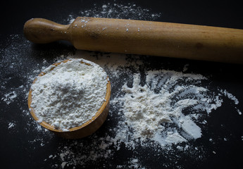 Bowl full of flour and rolling pin on a black table filled with flour