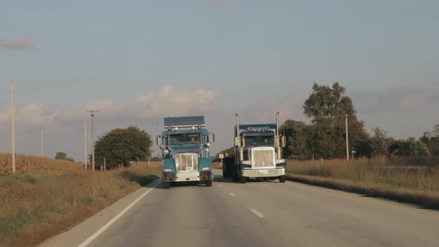 Two Peterbilt semi eighteen wheeler trucks driving down the highway in evening sun side by side in slow motion