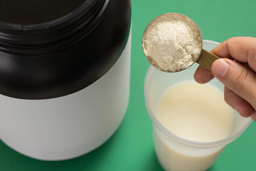 Whey Protein. Shaker with milk. Perspective, holding measuring scoop full of vanilla flavour powder on green background.