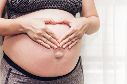 Portrait of pregnant woman making heart with hands on her belly near the window at home