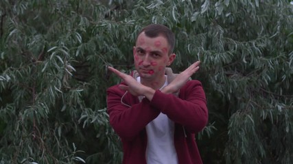 Young man with face full of lipstick marks of kisses shows stop sign gesture by crossing his hands
