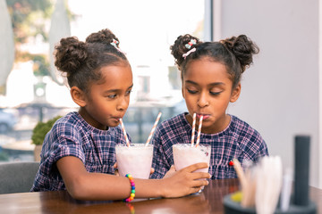 Share cocktail. Cute kind-hearted sisters wearing squared dresses sharing their milk cocktails with each other