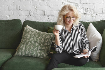 Fashionable elegant sixty year old lady with blonde hair and charming smile spending day indoors,...