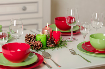 Merry Christmas and Happy New Year! Тable setting festive decor - green and red dishes, candles and fir cones. Knitted decor - Santa Claus knitted hats on the bottle with wine.
