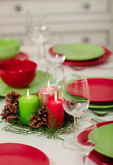 Merry Christmas and Happy New Year! Тable setting festive decor - green and red dishes, candles and fir cones. Knitted decor - Santa Claus knitted hats on the bottle with wine.