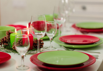 Merry Christmas and Happy New Year! Тable setting festive decor - green and red dishes, candles and fir cones. Knitted decor - Santa Claus knitted hats on the bottle with wine.