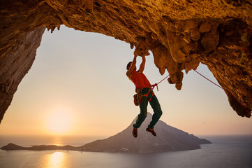 Male rock climber hanging on cliff with one hand at sunset