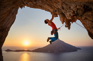 Male rock climber hanging with one hand on challenging route at sunset