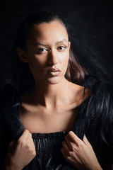 Low key close up portrait of pretty girl in fur vest and leather pants on dark background