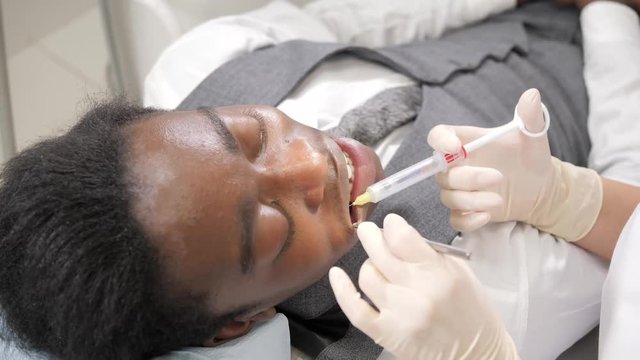 The dentist puts a shot of freezing novocaine in the jaw. Young African American male patient at chair at dental clinic. Medicine, health, stomatology concept.