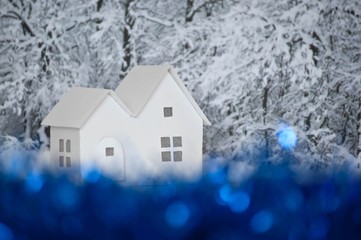 Fototapeta premium Small white cardboard houses, winter snowy trees background, blue bokeh effect. Handmade home winter decoration. Do it yourself. Shallow depth of focus.