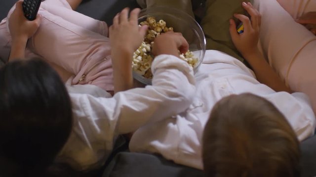 Top view of little boy and girl sitting on sofa, eating popcorn from glass bowl and holding remote control while watching TV at home