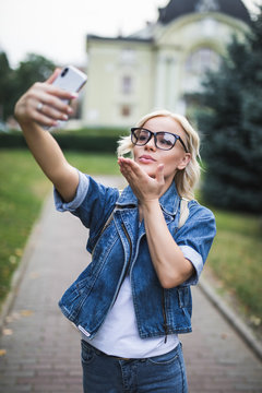 Beautiful blonde tourist woman hand holding smart phone photo camera lens, taking selfies pictures on holiday destination, blowing kisses, smiling looking outdoors