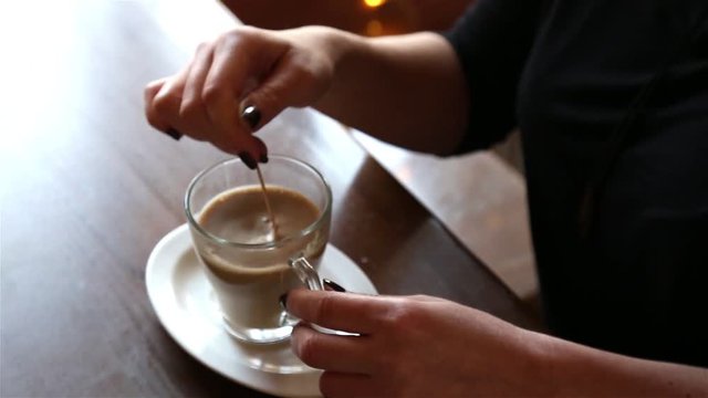 Woman mixes americano coffee in a glass cup sitting in the cafe at the wooden table. Hands close up, silhouette in backlight from the window.