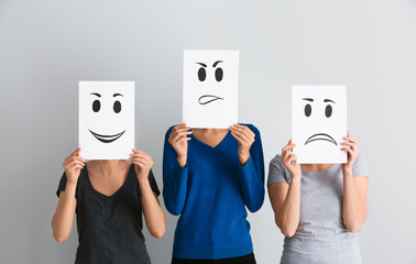 Young women hiding faces behind sheets of paper with drawn emoticons on light background