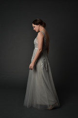 Obraz premium full length portrait of brunette girl wearing beautiful long gown, standing pose with background to the camera on grey studio background.