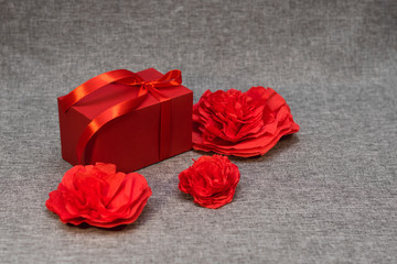 the gift box is wrapped in a red ribbon, and with paper hearts and flowers around, on a gray background