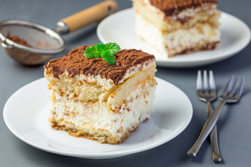 Two piece of traditional italian Tiramisu dessert cake on a white plate, decorated with cocoa powder and mint, on gray background, horizontal