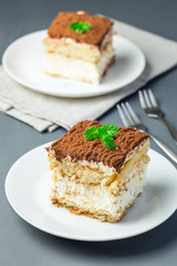 Two piece of traditional italian Tiramisu dessert cake on a white plate, decorated with cocoa powder and mint, on gray background, vertical