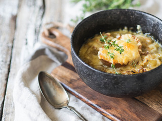 Traditional french onion soup with toast, cheese and fresh thyme in a black bowl on a wooden brown board on a rustic wooden background. Copy space
