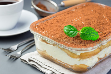 Traditional italian Tiramisu dessert cake in a glass form, decorated with cocoa powder and mint, with coffee cup, on gray background, horizontal