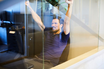 Creative businessman sitting with arms raised seen through glass in office