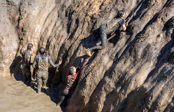 Young people pass the obstacle course. Mud race runners. All in the mud. Deep dirty pit, climb the rope.