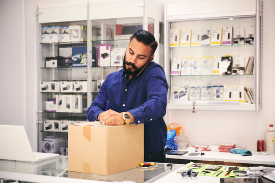 Male owner talking on mobile phone while packing box at counter in electronics store